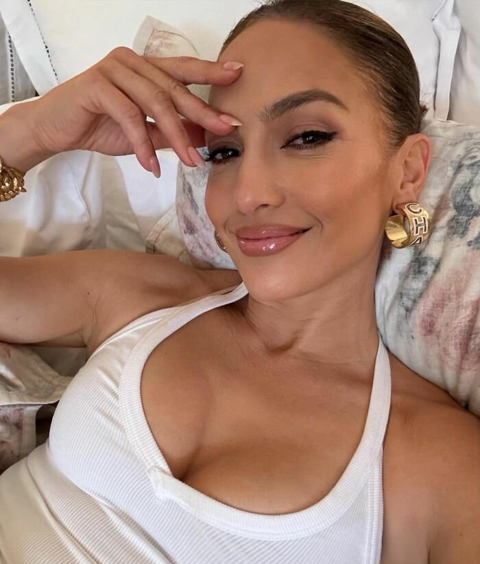 Jennifer Lopez smiling in a white tank top wearing gold CH earrings with a relaxed pose indoors.