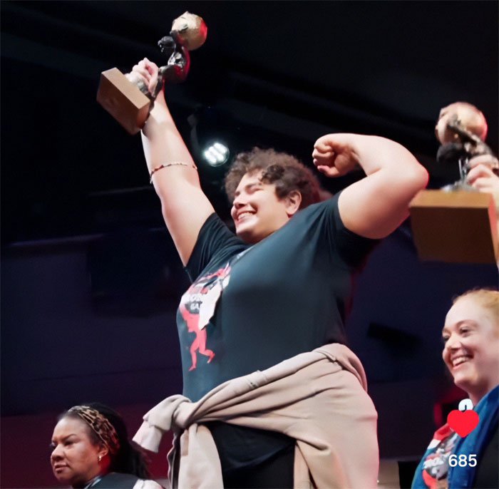 Woman holding trophy and celebrating on stage at strongwoman competition, highlighting world&rsquo;s strongest woman controversy.