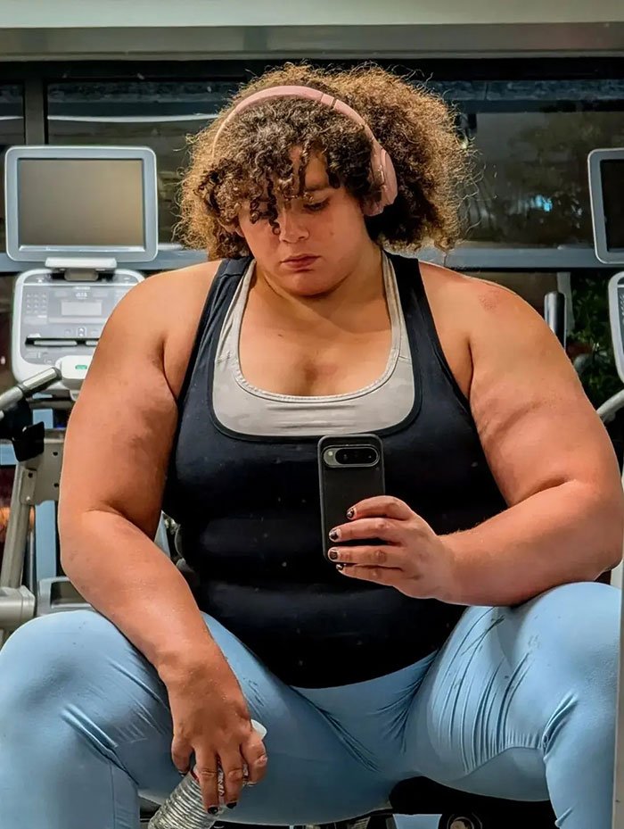 Strong woman with muscular arms wearing headphones and workout clothes, taking a gym selfie sitting on a bench.
