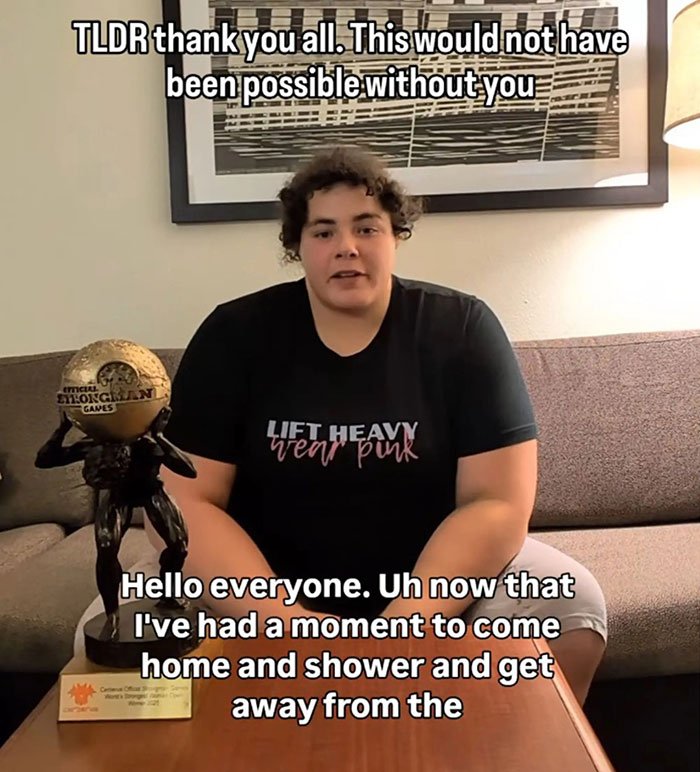 Person sitting on a couch beside a strongman trophy, discussing the world's strongest woman and related controversy.