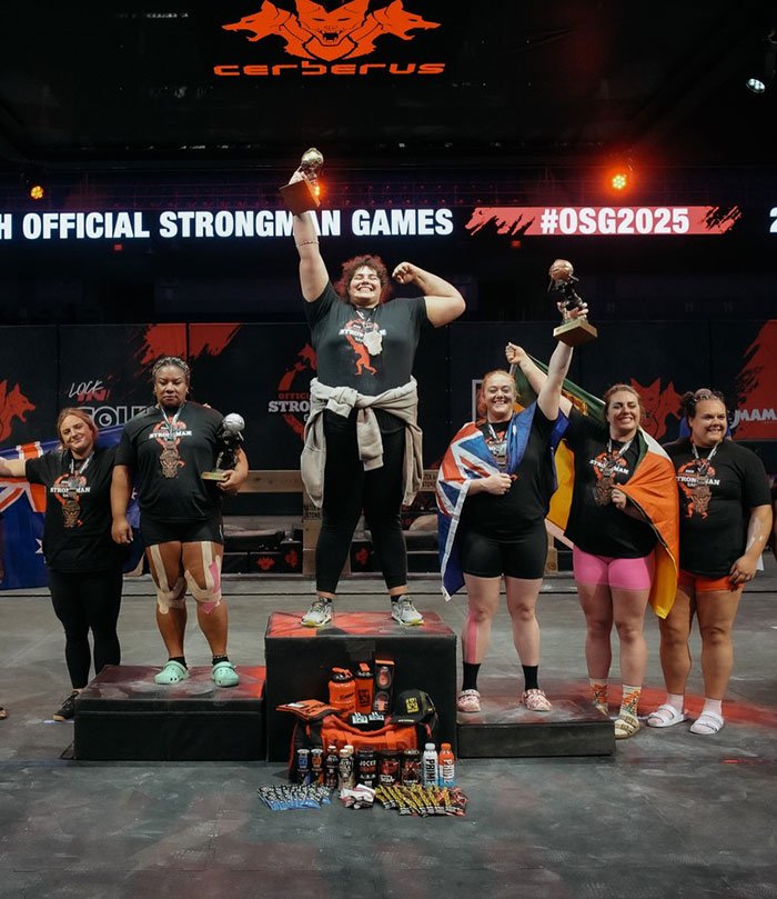Podium scene with strongwoman athletes celebrating at official strongman games event with trophies and medals.