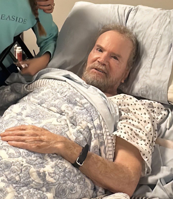 Elderly gravely ill veteran lying in hospital bed, covered with blanket, receiving care from a person nearby.