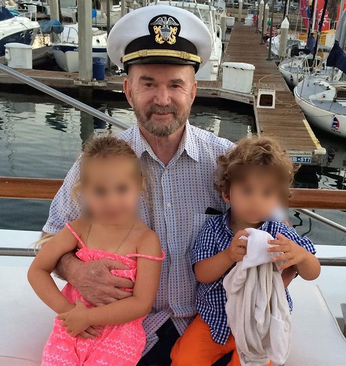 Elderly veteran in a naval officer hat smiling and holding two young children on a boat docked at a marina.