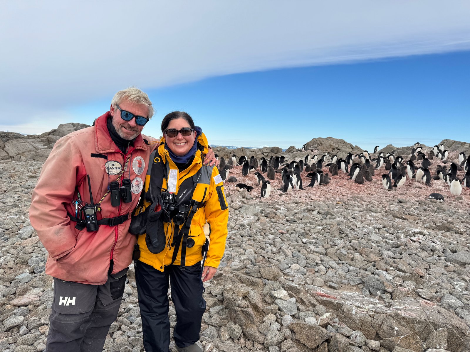 Two explorers in cold weather gear smiling on rocky land with a large group of penguins in Antarctica’s stunning landscape.