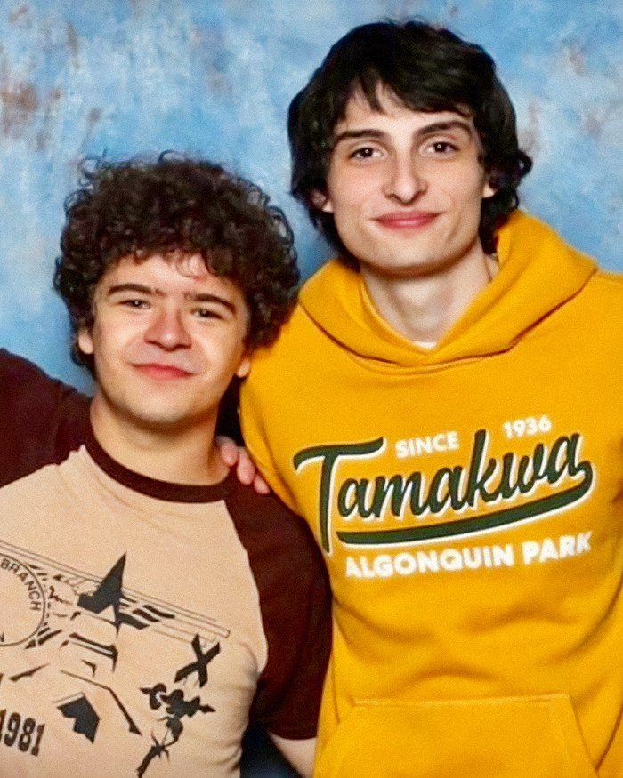 Stranger Things actor Gaten Matarazzo and co-star smiling together against a blue backdrop in casual clothing.