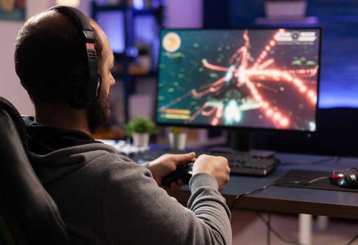 Man wearing headphones playing video games on computer, avoiding Thanksgiving shopping pressure from wife’s ban decision.