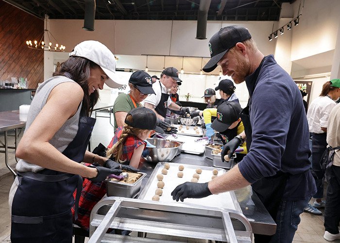 Meghan Markle participating in a volunteer event, involved in an awkward royal gesture with others preparing food.