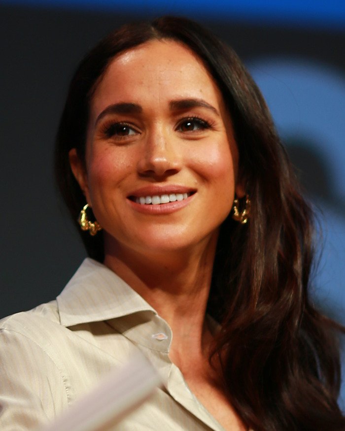 Meghan Markle smiling during an event, wearing gold earrings and a cream shirt with an awkward royal gesture discussed.