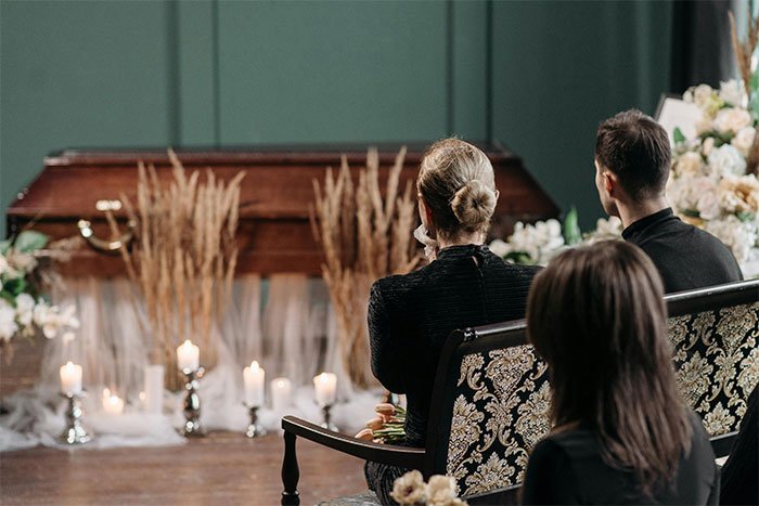 Three mourners sitting at a funeral near a coffin, symbolizing family protecting daughter from solicitor about inheritance.