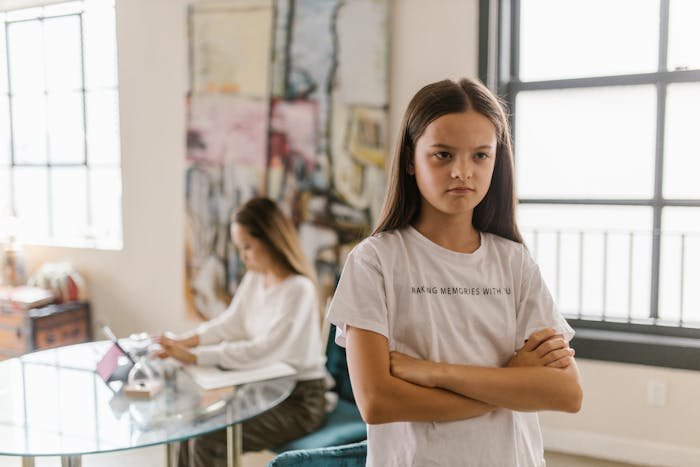 Young girl with folded arms looking upset while mom works at table, depicting mom doesn't like how her kids act issues.