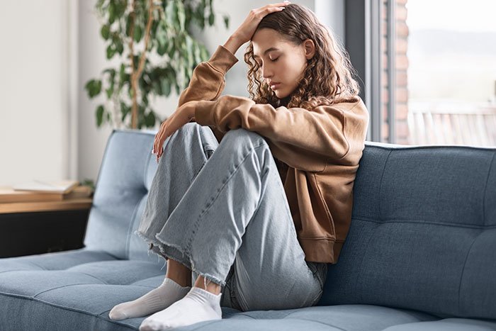 Young woman sitting on couch with knees drawn up, appearing upset after selfish dad demands apology from daughter.