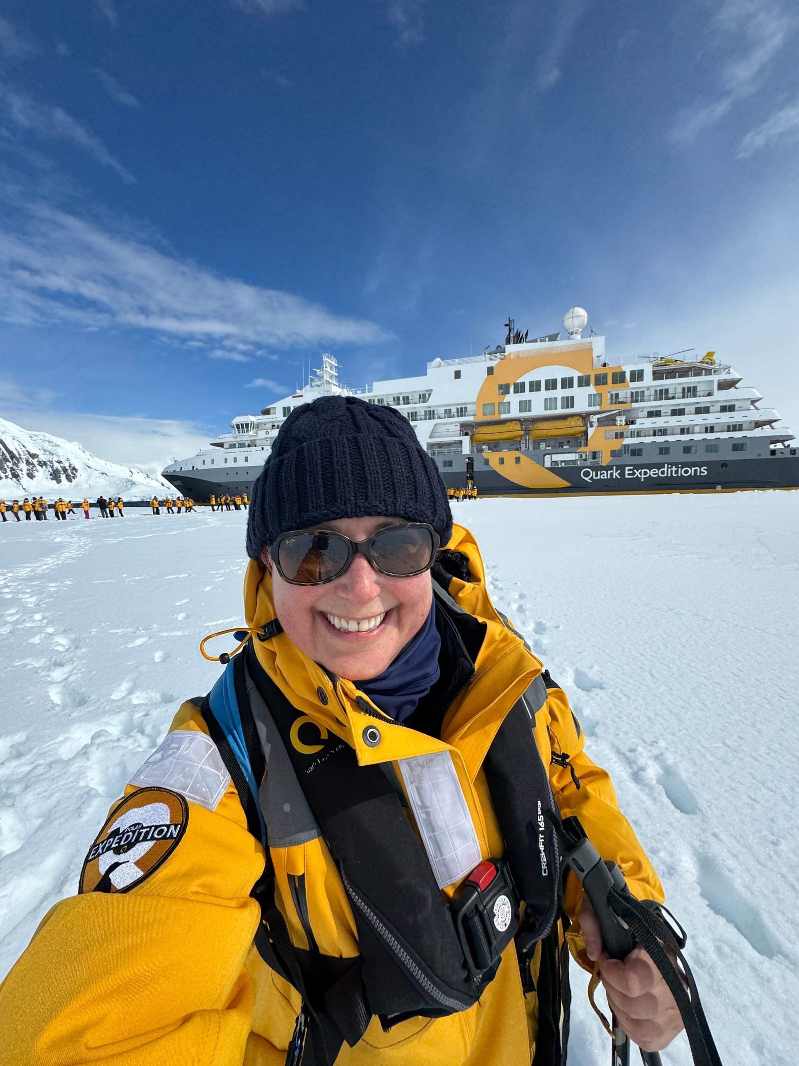 Smiling explorer in yellow gear taking a selfie on the snow with an Antarctica expedition ship and group in the background.