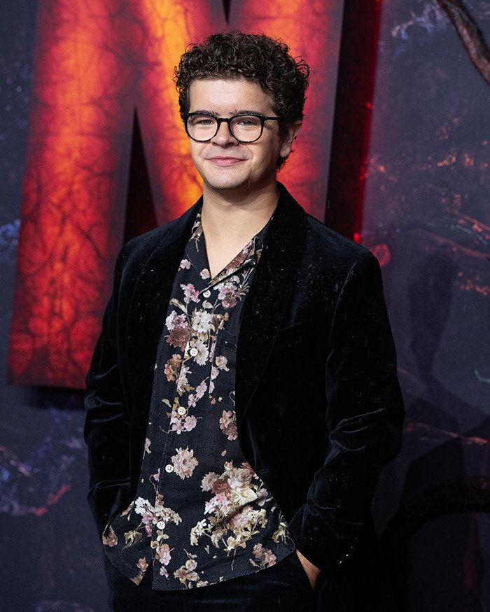 Stranger Things actor Gaten Matarazzo wearing glasses and floral shirt at a red carpet event with illuminated backdrop.