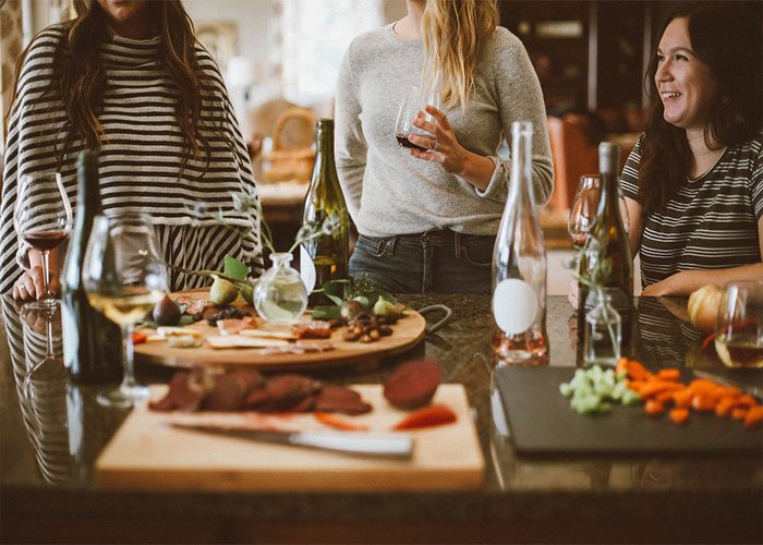 Three women chatting and drinking wine around a kitchen island with snacks, related to birthday parties and texts revealed.