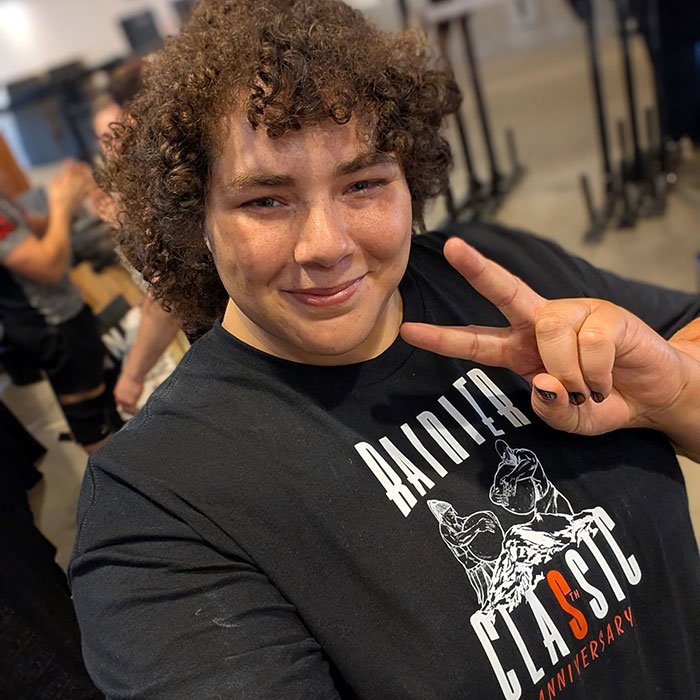 Person with curly hair wearing a black shirt and making a peace sign, linked to world&rsquo;s strongest woman controversy.