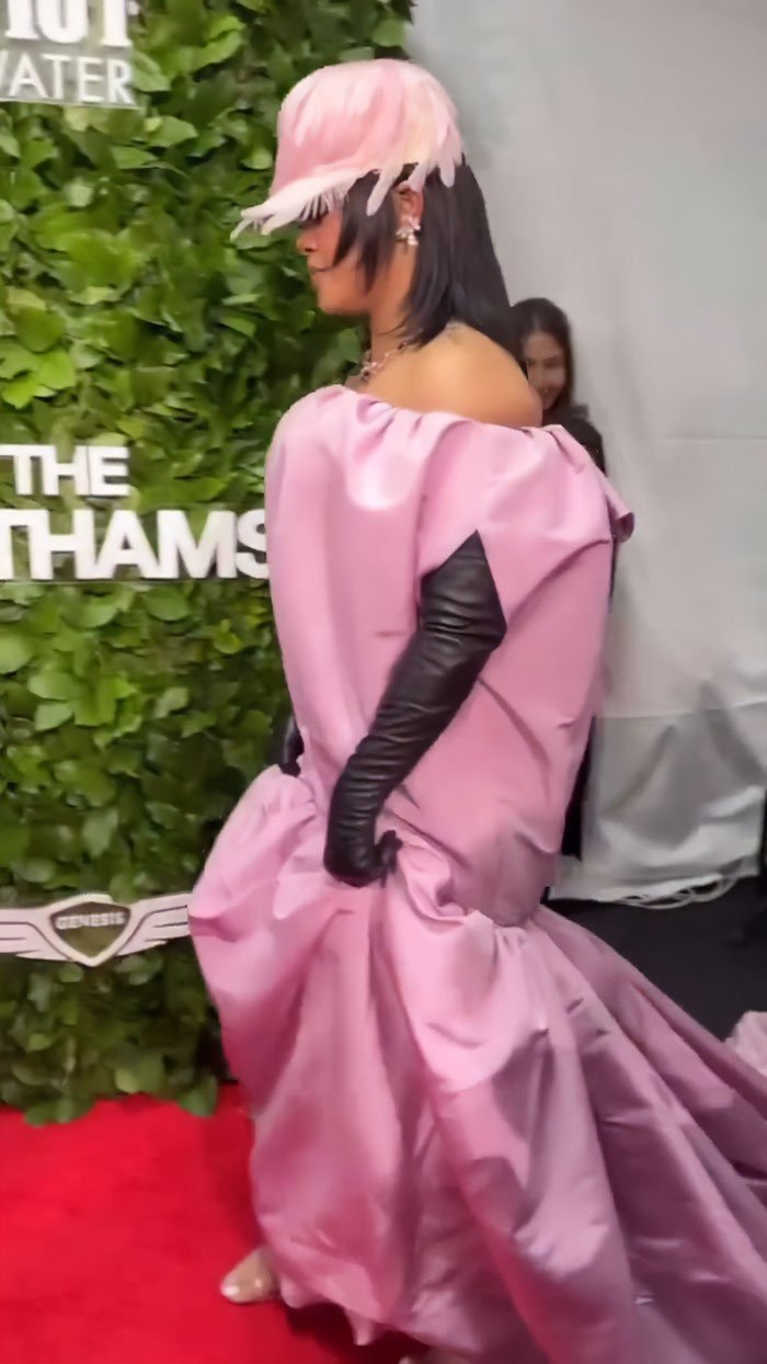 Rihanna wearing a voluminous pink gown with black gloves and a matching pink feathered hat at a red carpet event.