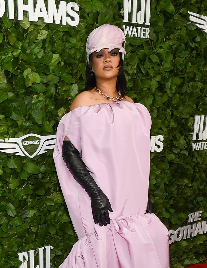 Rihanna wearing a voluminous pink dress and matching hat with black gloves on a green leafy red carpet backdrop