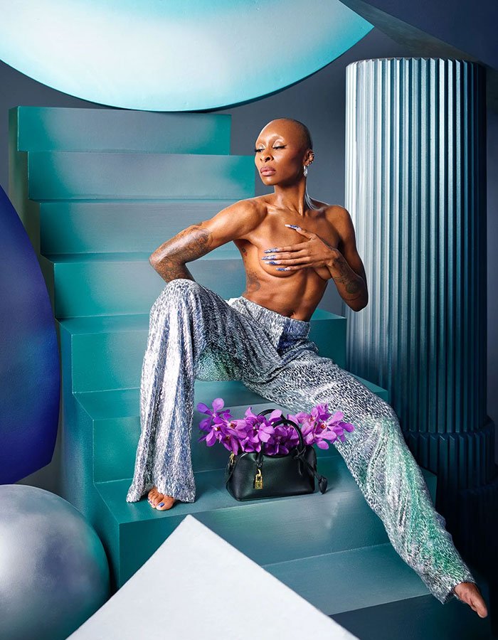 Cynthia Erivo posing topless on teal stairs with silver pants and a handbag filled with purple flowers in artistic setting
