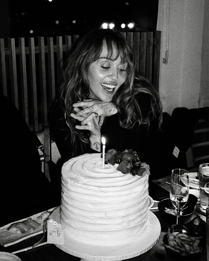Miley Cyrus smiling at a cake with a candle, celebrating with engagement and mixed reactions about age.