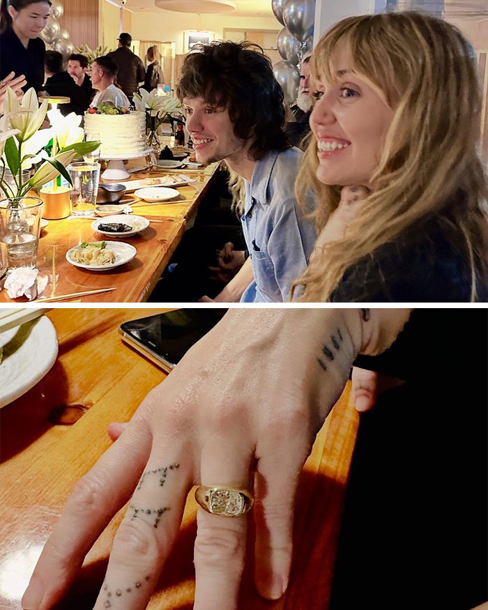 Miley Cyrus and Maxx Morando smiling at engagement party with close-up of gold ring on finger at wooden table.