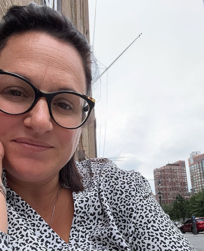 Woman wearing glasses outdoors in a city setting, reflecting on emotional experience related to body-shaming on flight.