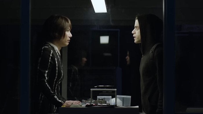 BD Wong from Jurassic World in a tense conversation wearing a striped outfit facing a person in a black hoodie indoors.