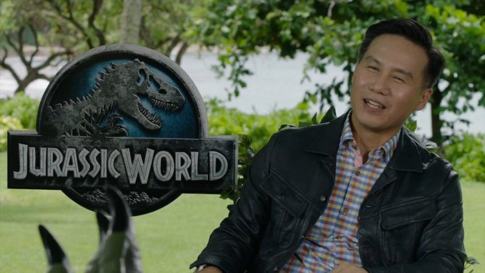 BD Wong from Jurassic World outdoors near a Jurassic World sign, wearing a black jacket and plaid shirt.
