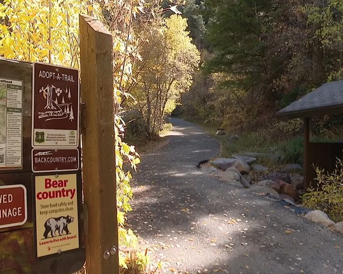 Mountain trail entrance with signs warning about bear country and safety tips along a forest path in autumn foliage.