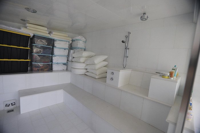 Sterile tiled bathroom inside Epstein’s private island home with shower and stacked pillows and storage containers.