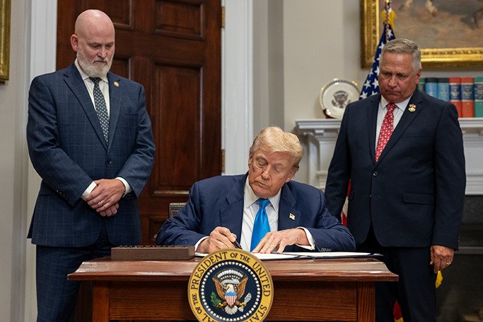 Former President signing documents in an official setting with two men standing nearby, symbolizing Epstein’s private island home reveal.