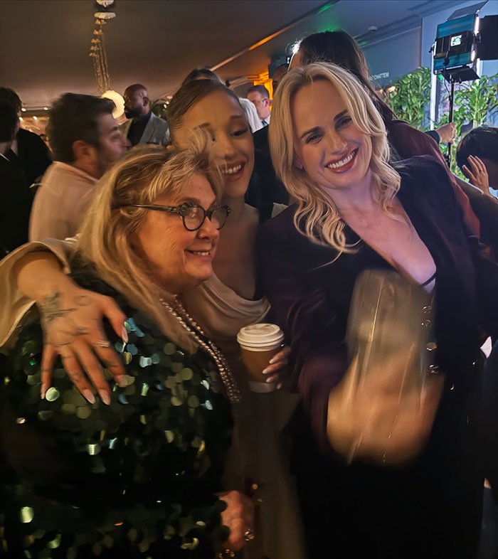 Rebel Wilson and Ariana Grande smiling and posing for a selfie at a crowded indoor event with people in the background.