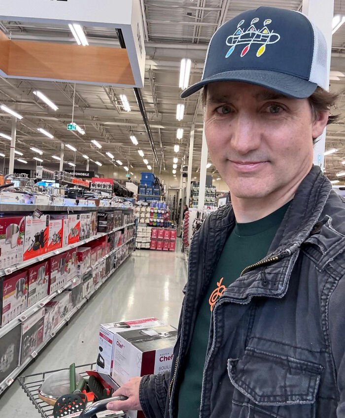 Justin Trudeau shopping in a store aisle, casually dressed with a cap, holding a cart with kitchen appliances.