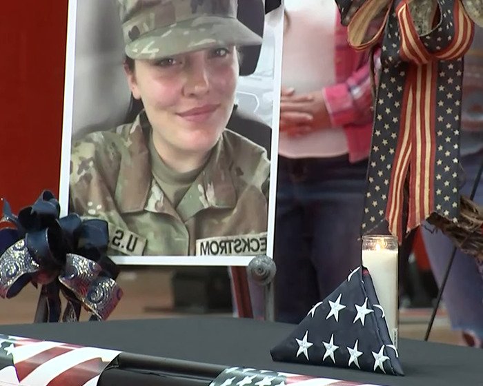 Memorial setup with a military photo, folded American flags, patriotic ribbons, and a lit white candle on a table.
