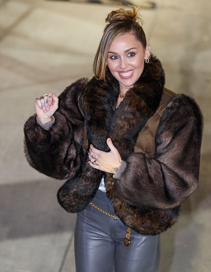 Miley Cyrus smiling and posing outdoors in a fur coat, showcasing her youthful glow and viral makeover.