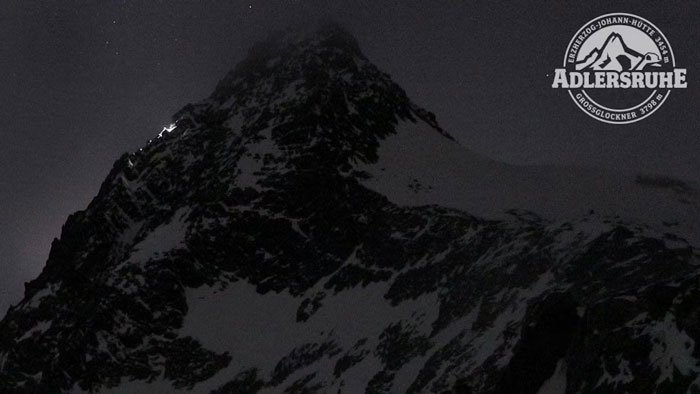 Snowy peak of Austria’s highest mountain at night with climbers' lights and Adlersruhe emblem in the sky corner.