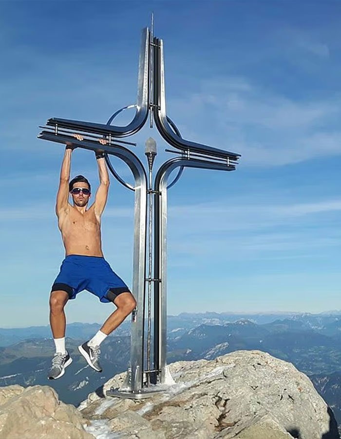 Man wearing sunglasses and blue shorts hanging from metal cross on Austria’s highest peak with mountain landscape behind him