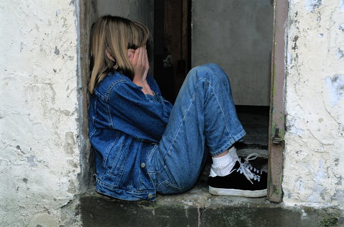 Young girl in denim jacket sitting on doorstep covering her face, reflecting a daughter doing much for her mother.