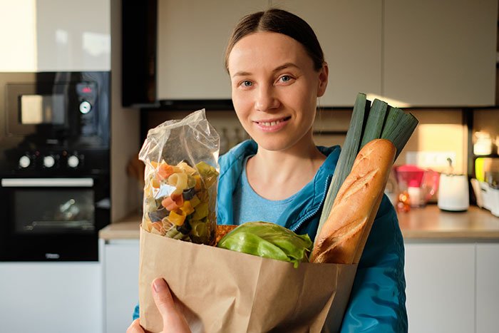 Woman holding groceries in kitchen, representing mom accused of embarrassing son’s girlfriend who offered to cook meals.
