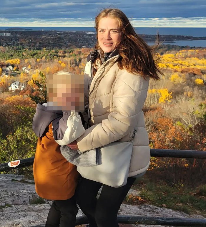 Woman in beige jacket holding child with blurred face outdoors in autumn landscape related to racist Cinnabon worker news.