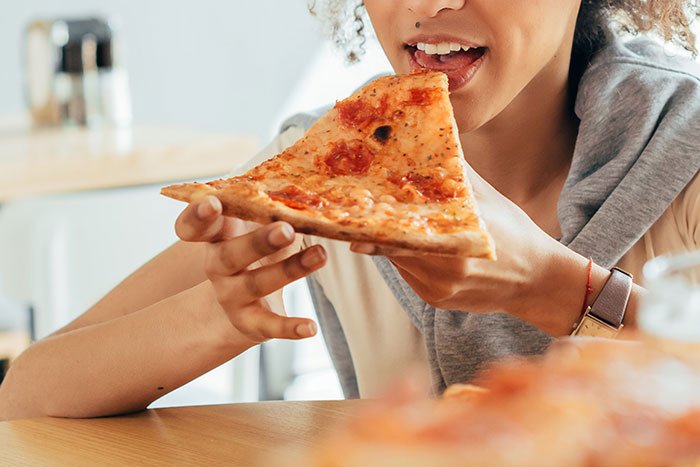 Young babysitter eating a slice of pizza at a table as parents look on puzzled by her confession of having two slices.