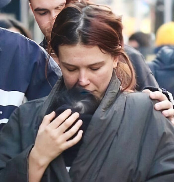 Millie Bobby Brown tenderly holding newborn daughter wrapped in dark clothing amidst a busy outdoor crowd.