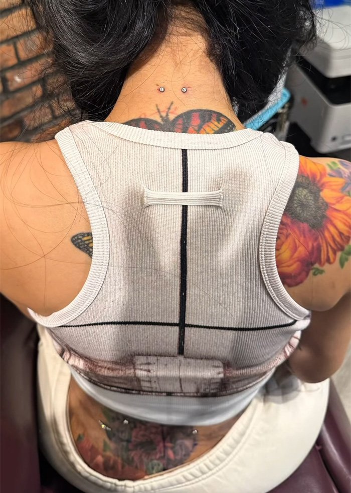 Close-up of neck and back piercings with colorful butterfly and flower tattoos on skin beneath white tank top.