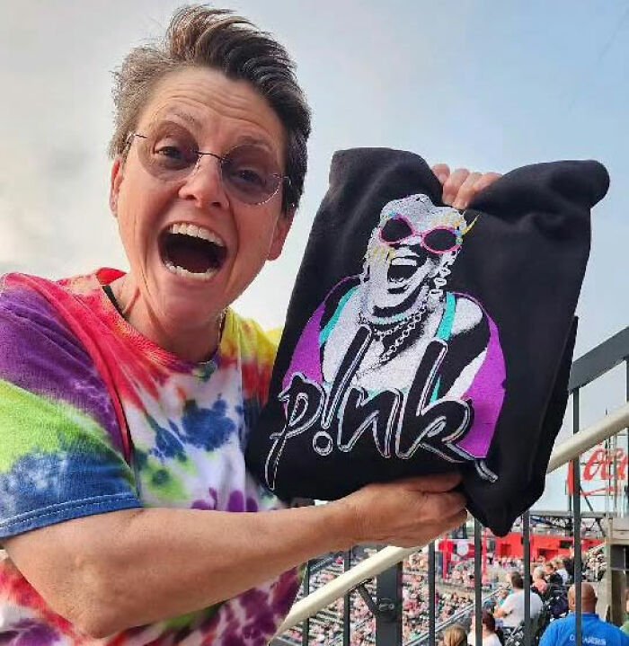 Actress from Marvelous Mrs. Maisel wearing glasses and tie-dye shirt, holding a colorful P!nk sweatshirt outdoors.