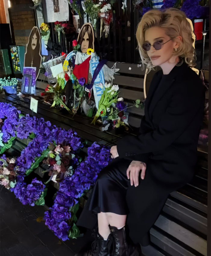 Woman with sunglasses and blonde hair sitting by a memorial adorned with flowers, related to Sharon Osbourne and Kelly&rsquo;s weight loss news.