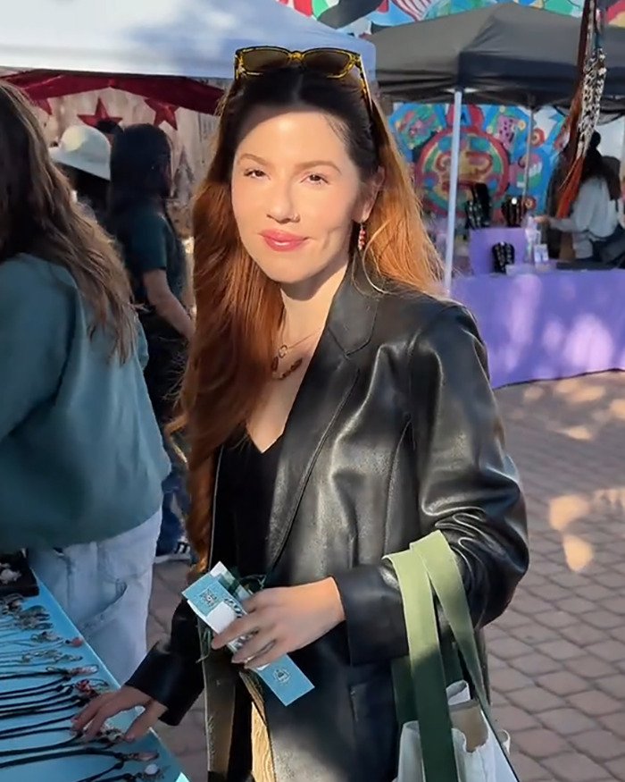 Young woman at outdoor market smiling, wearing black leather jacket and sunglasses, related to woke social media user topic.