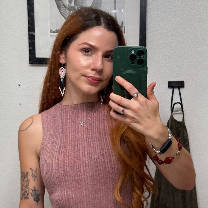 Young woman taking a selfie while wearing a pink sleeveless top, showcasing tattoos and long reddish hair for social media post.