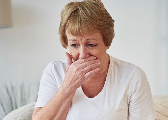 Upset woman covering mouth with hand, reacting emotionally indoors to her son’s partner and gift expectations.