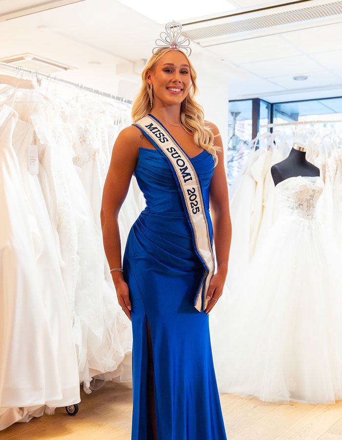 Miss Finland wearing blue gown and crown, posing indoors among wedding dresses before losing crown amid social media backlash.