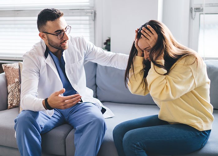 Worried woman holding head as man in white coat talks, depicting conflict over future child&rsquo;s health risk and exposed lie.