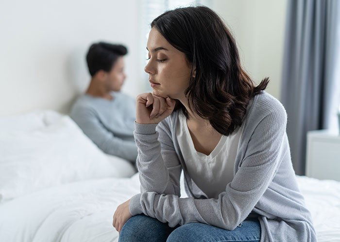 Woman looks upset sitting on bed while man in background appears distant, highlighting future child health concerns after a lie exposed.